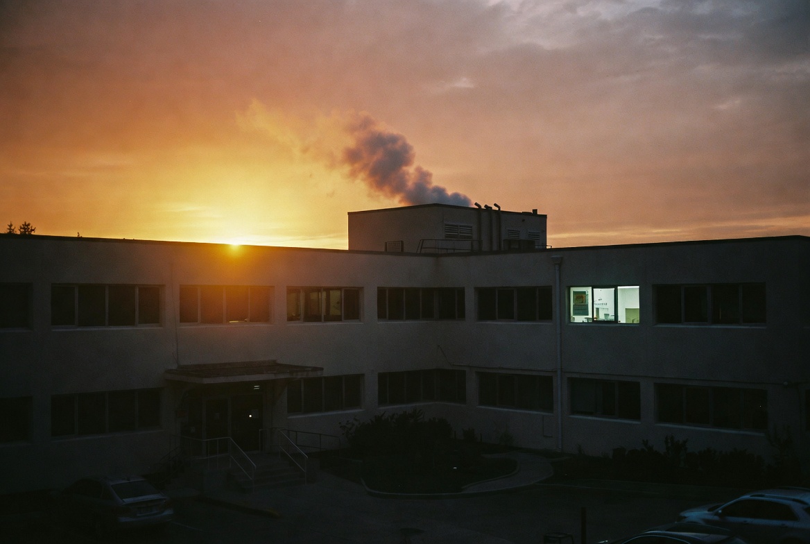 The office building at sunset, smoke rising from the roof vent, one office window still lit against the fading sky
